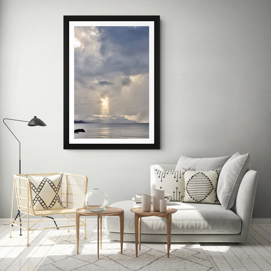 A framed vertical art print depicting a dramatic cloudy sky with sunbeams over a calm sea, styled in a modern living room setting above a light gray sofa.