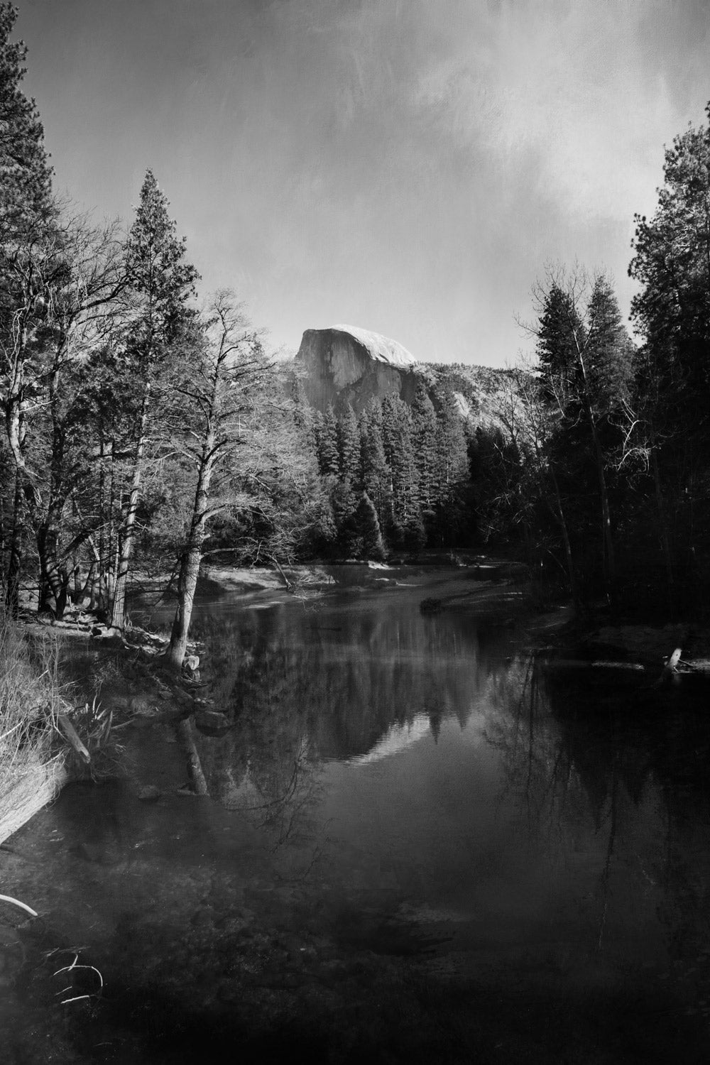 Black and white photograph of Half Dome mountain in Yosemite National Park, reflecting in a still lake surrounded by trees.